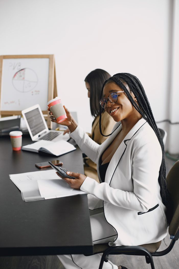 Smiling woman in office attire holding a coffee cup and using a calculator in a modern office.